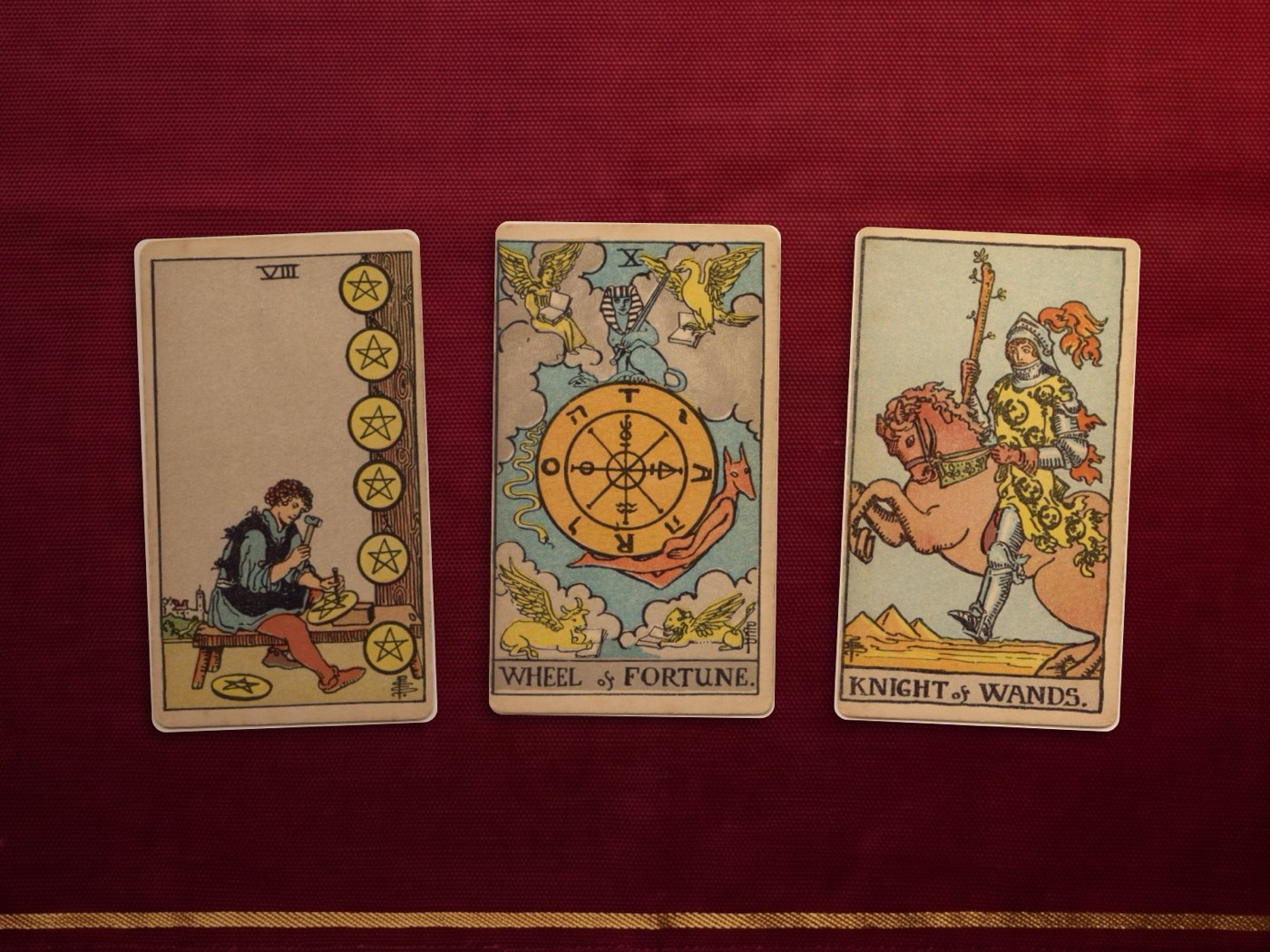 Three tarot cards on a maroon cloth: the Eight of Pentacles, the Wheel of Fortune and the Knight of Wands.